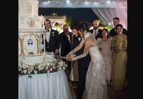 Newly weds Nick Jonas and Priyanka Chopra can be seen cutting their 7-tier wedding cake with a sword-like knife as their friends and family watch on. (Photo | Instagram)