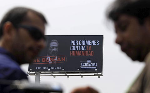 Pedestrians walk below a billboard showing an image of Cuba's President Raul Castro and a message that reads in Spanish: 'Wanted for crimes against humanity' | AP