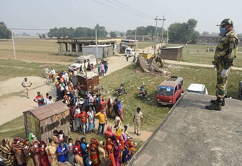 A security personnel keeps vigil as voters stand in long queues to cast their votes at a polling station. (Photo | PTI)