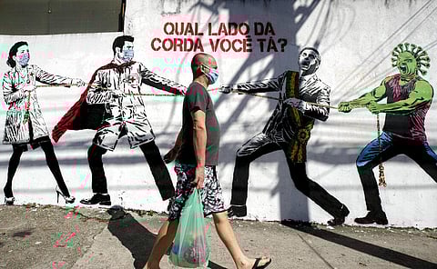 A man, wearing a protective face mask as a measure to curb the spread of the new coronavirus, walks past a mural depicting a tug-of-war between health workers and Brazil's President Jair Bolsonaro aided by a cartoon-styled coronavirus character, with a me