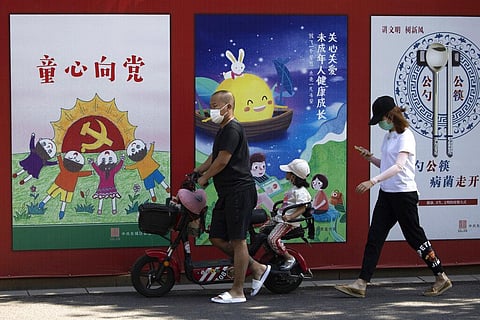 A man wearing a mask to protect from the coronavirus pushes an electric bike with a child past propaganda posters with the words 'Childlike devotion to the Party' in Beijing. (Photo | AP)