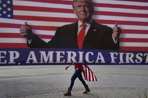 a woman draped in an American flag walks past a banner supporting President Donald Trump during a rally in Huntington Beach, California. (Photo | AP)