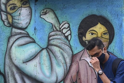 A commuter adjusts his mask while walking past a wall mural depicting frontline warriors of the Covid-19. (Photo| AFP)