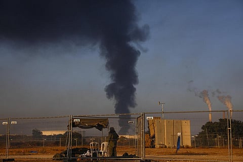 An Israeli soldier stands guard next to an Iron Dome air defense system as smoke rises from an oil tank on fire after it was hit by a rocket fired from Gaza Strip, near Ashkelon. (Photo | AP)