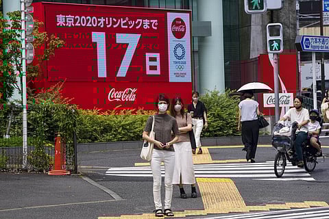 People wearing protective masks wait at a crosswalk as an electric display in the background shows 17 days to Tokyo 2020 Olympics. (Photo | AP)