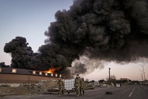 Ukrainian servicemen carry containers backdropped by a blaze at a warehouse after a bombing on the outskirts of Kyiv, Ukraine. (Photo | AP)