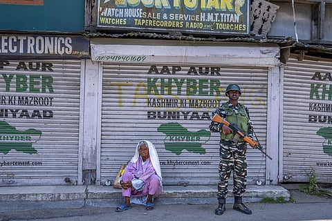 A paramilitary soldier stands guard beside a woman resting outside closed shops during shutdown in central Srinagar. (Photo | AP)