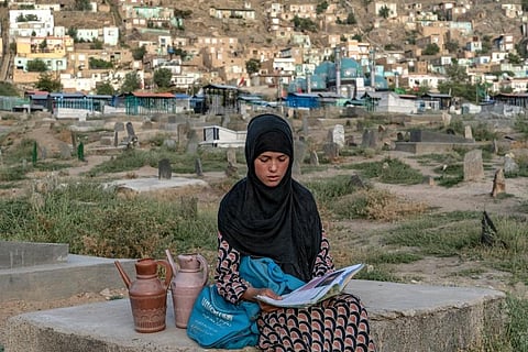 Sara, a 14-year-old, an Afghan girl, sits on a grave and reads a book as she waits for customers to sell water at a cemetery, in Kabul, Afghanistan. (Photo | AP)