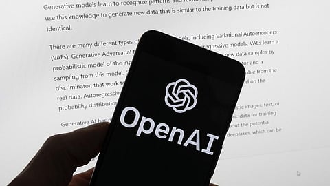 The OpenAI logo is seen on a mobile phone in front of a computer screen displaying output from ChatGPT