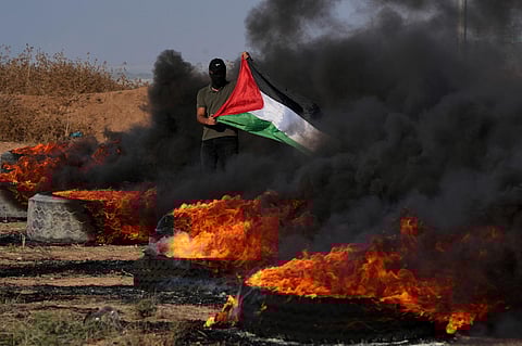 A masked Palestinian protester waves his national flag next to burning tires during clashes with Israeli security forces along the frontier with Israel. (Photo | AP)