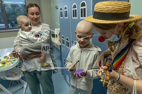Tetiana Nosova, who goes by the clown name of Zhuzha, a volunteer from the "Bureau of Smiles and Support" plays a ukulele as she stands with Michael Bilyk, who is held by his mother Antonina Malyshko, and Kira Vertetska, 8, at Okhmatdyt children's hospital in Kyiv, Ukraine, Thursday Sept. 19, 2024. (AP Photo/Anton Shtuka)