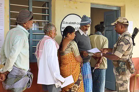 A security personnel checks voter ID as voters wait to cast votes at a polling station during the Channapatna Assembly by-poll, Karnataka