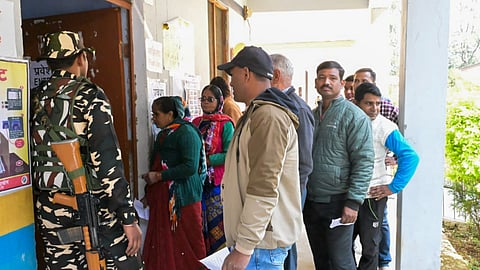 A security personnel stands guard as voters wait in queues to cast votes at a polling booth during the Kedarnath Assembly constituency bypoll.