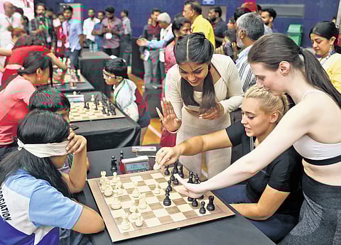 A Miss World contestant, flanked by her counterparts from other countries offering tips, plays chess with a blindfolded girl