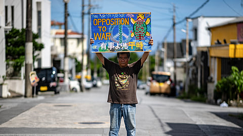 This photo taken on June 8, 2025, shows Takemasa Kinjo, who was a high school student when his mother was killed by a US Marine in 1974, holding a placard reading "Opposition to War" outside his residence in the Henoko area of Nago city, Okinawa Prefecture.