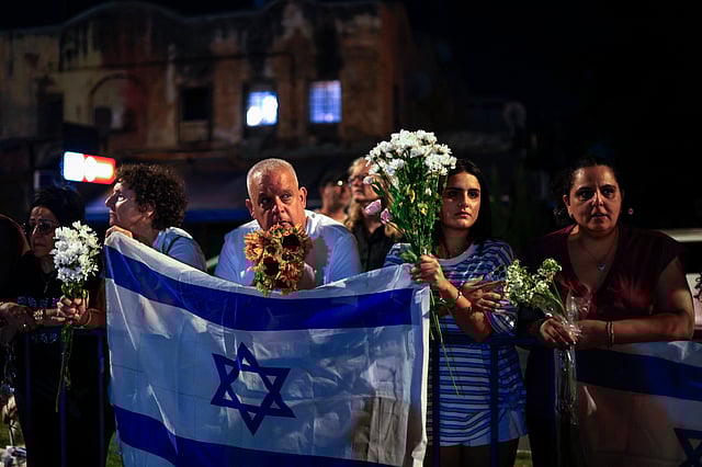 People carry flowers and Israeli flags upon the arrival of vehicles transporting the bodies of four hostages handed over following a ceasefire and prisoner exchange deal between Israel and Palestinian factions in Gaza, in front of the National Center for Forensic Medicine in Tel Aviv on October 13, 2025.