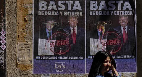 A woman waits for the bus in front of a poster with an image of President Javier Milei and President Donald Trump and a message that reads in Spanish; "Enough of debt and surrender'" in Buenos Aires, Argentina, Thursday, Oct. 2, 2025.