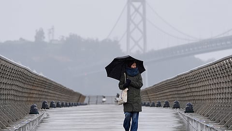 A woman walks in the rain as the San Francisco-Oakland Bay Bridge is seen in the background, Tuesday, Feb. 17, 2026, in San Francisco.