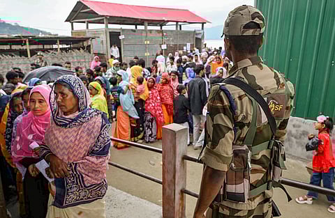 A security personnel stands guard as people wait in queues to cast their votes during the Assam Assembly elections, at a polling station, in Darrang district, Assam, Thursday, April 9, 2026.