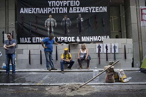 Protesting hospital staff sit in front of a wall that they built at the entrance of the Greek Finance Ministry with a banner depicting Greek Prime Minister Alexis Thipras , Deputy Health Minister Pavlos Polakis and Greek Finance Minister Euclid Tsakalotos