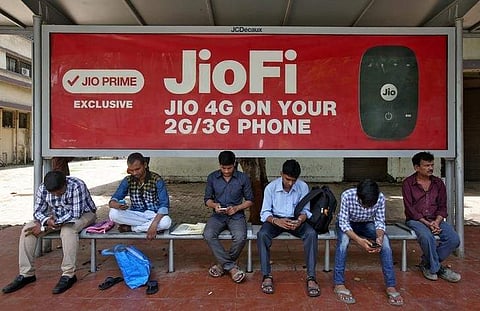 Commuters use their mobile phones as they wait at a bus stop with an advertisement of Reliance Industries' Jio telecoms unit in Mumbai.(Photo | Reuters)
