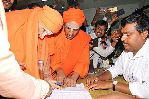 Centenarian Sri Shivakumara Swamiji just before casting his vote puts his signature at a polling station at Siddaganga Mutt in Tumakuru rural assembly segment on Saturday. His junior Sri Siddalinga Swamiji helps him. | EPS