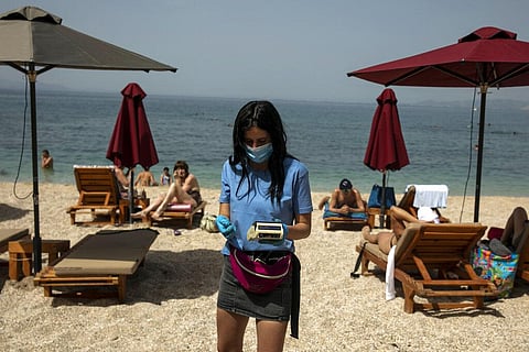 An employee of a beach bar wearing a protective mask against coronavirus, collects money for sunbeds from the swimmers, at Alimos beach, near Athens. (Photo | AP)