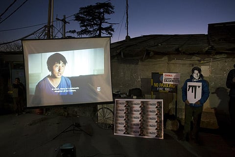 An exile Tibetan wearing a mask of 32-year-old Tashi Wangchuk, stands next to a screen projecting a New York Times video during a street protest demanding his release, in India. (Photo | AP)