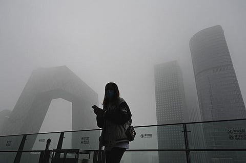 A woman uses her phone along a glass wall with the China Central Television (CCTV) headquarters in the background on a polluted day in Beijing  (Photo | AFP)