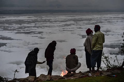 People warm themselves in front of small fire on the banks of Yamuna river as toxic foam floats on the surface at Kalindi Kunj. (Photo | PTI)
