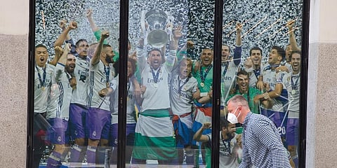 A masked man and a young boy walk past a Real Madrid poster of the team celebrating in a merchandising shop in Madrid, Spain. (File Photo | AP)
