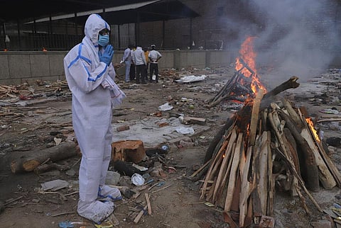 A man wearing personal protective equipment prays in front of the burning funeral pyre of his father who died of COVID-19, at a crematorium in New Delhi. (Photo | AP)