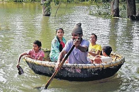 Mujahid Pasha, who runs a chicken stall, helps people cross a flooded layout on his coracle, free of cost, in Srirangapatna on Saturday | Udayashankar S