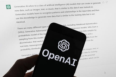 The OpenAI logo is seen on a mobile phone in front of a computer screen displaying output from ChatGPT. (Photo | AP)