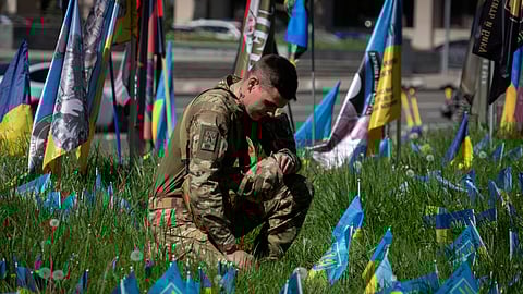 A soldier stands kneeling at the Independence square in Kyiv, Ukraine, Wednesday, May 1, 2024, surrounded by small national flags symbolising Ukrainian soldiers killed in the war with Russia.
