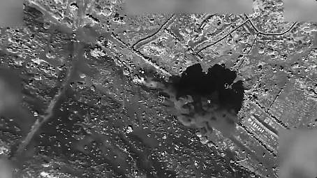 The footage cuts to a wider shot showing a column of smoke over the apparent impact site and several vehicles parked further up the road.