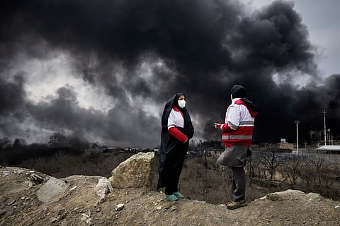 Two women from the Iranian Red Crescent Society stand as a thick plume of smoke from a U.S.-Israeli strike on an oil storage facility late Saturday rises in the sky in Tehran, Iran, Sunday, March 8, 2026.