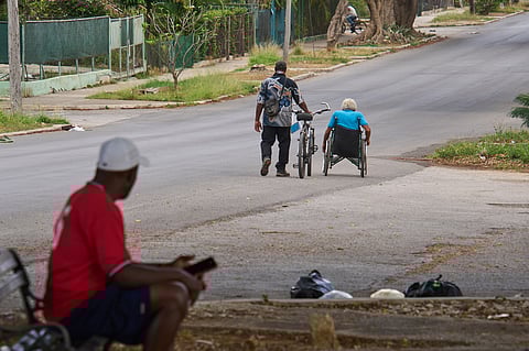 An elderly man makes his way in his wheelchair while a friend walks a bicycle beside him, in Havana, Cuba, Wednesday, April 15, 2026.